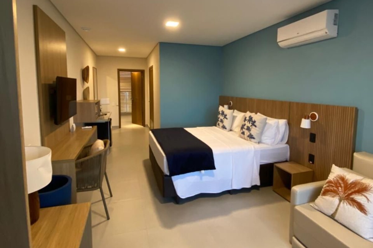 A modern hotel room with a bed, a desk, a chair, a sofa, and an air conditioning unit.