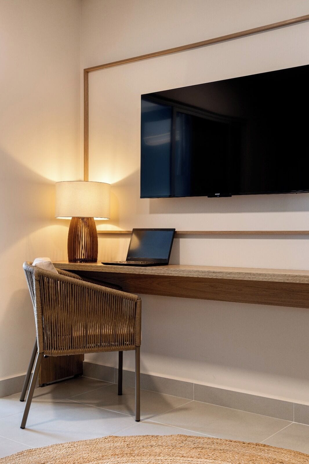 Quarto moderno com televisão de tela plana na parede, um laptop sobre uma escrivaninha de madeira, uma cadeira de vime e um abajur.