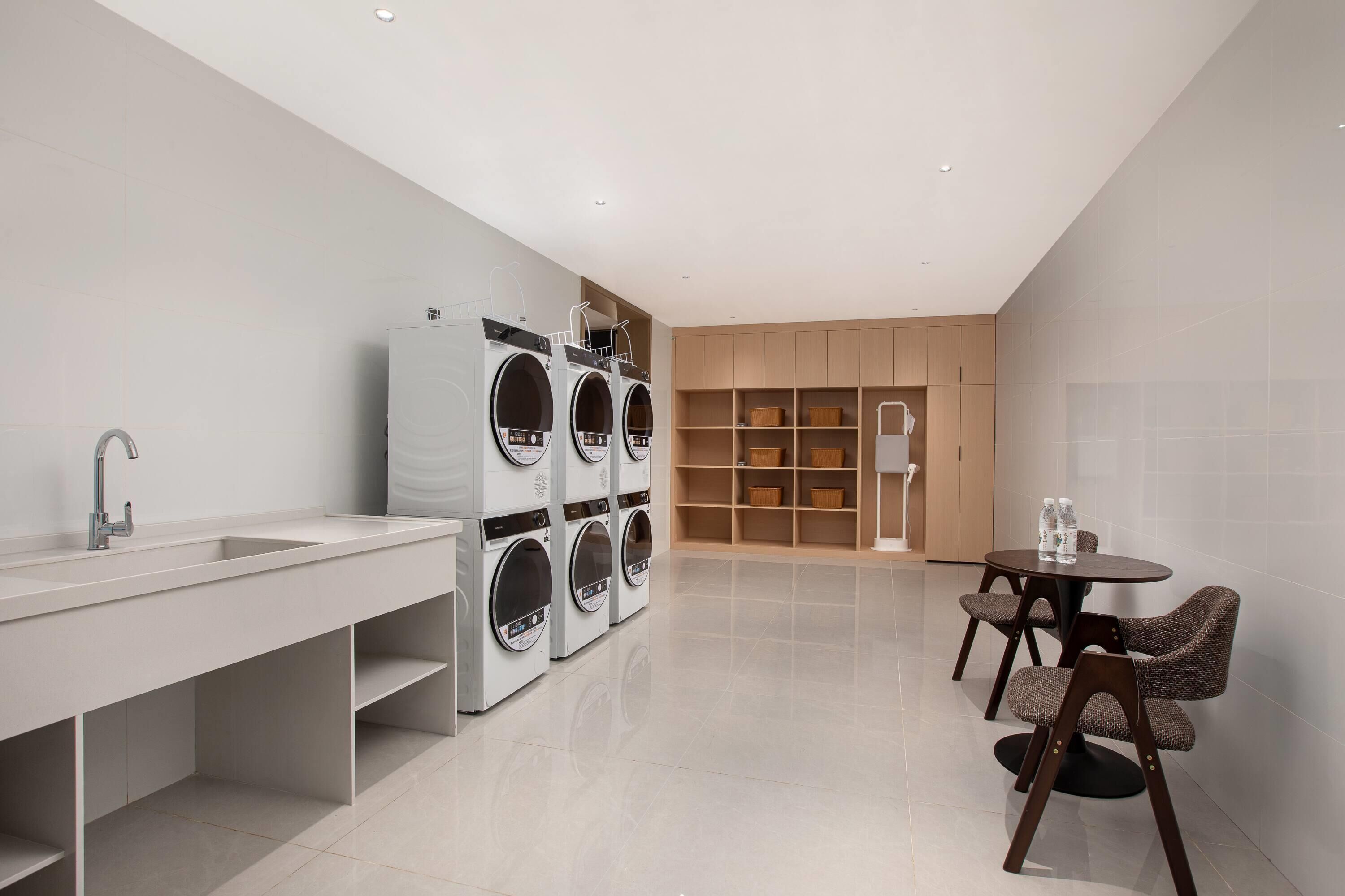 A modern bathroom with a large sink, a mirror, and a set of stacked washing machines. There is a round table with chairs and a small lamp.