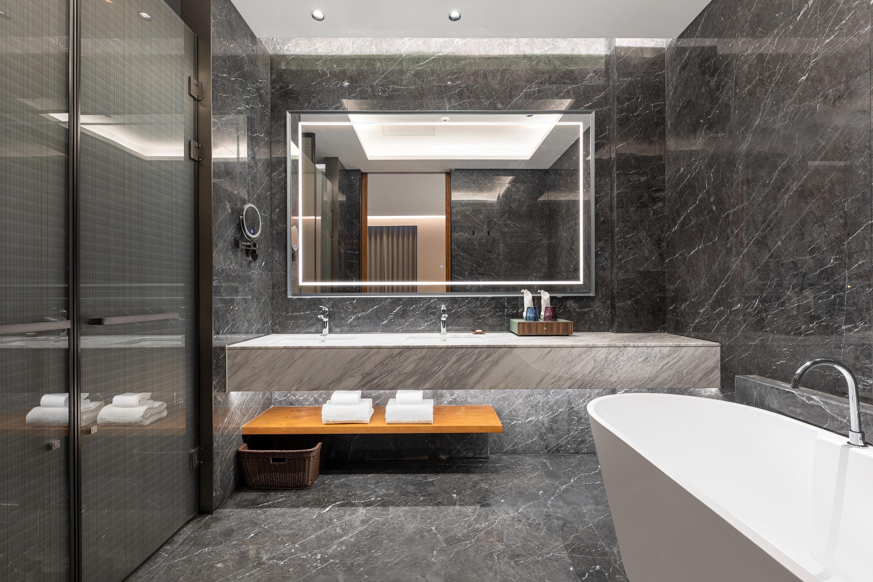 A modern bathroom with a large mirror, two sinks, a bathtub, and a wooden bench.
