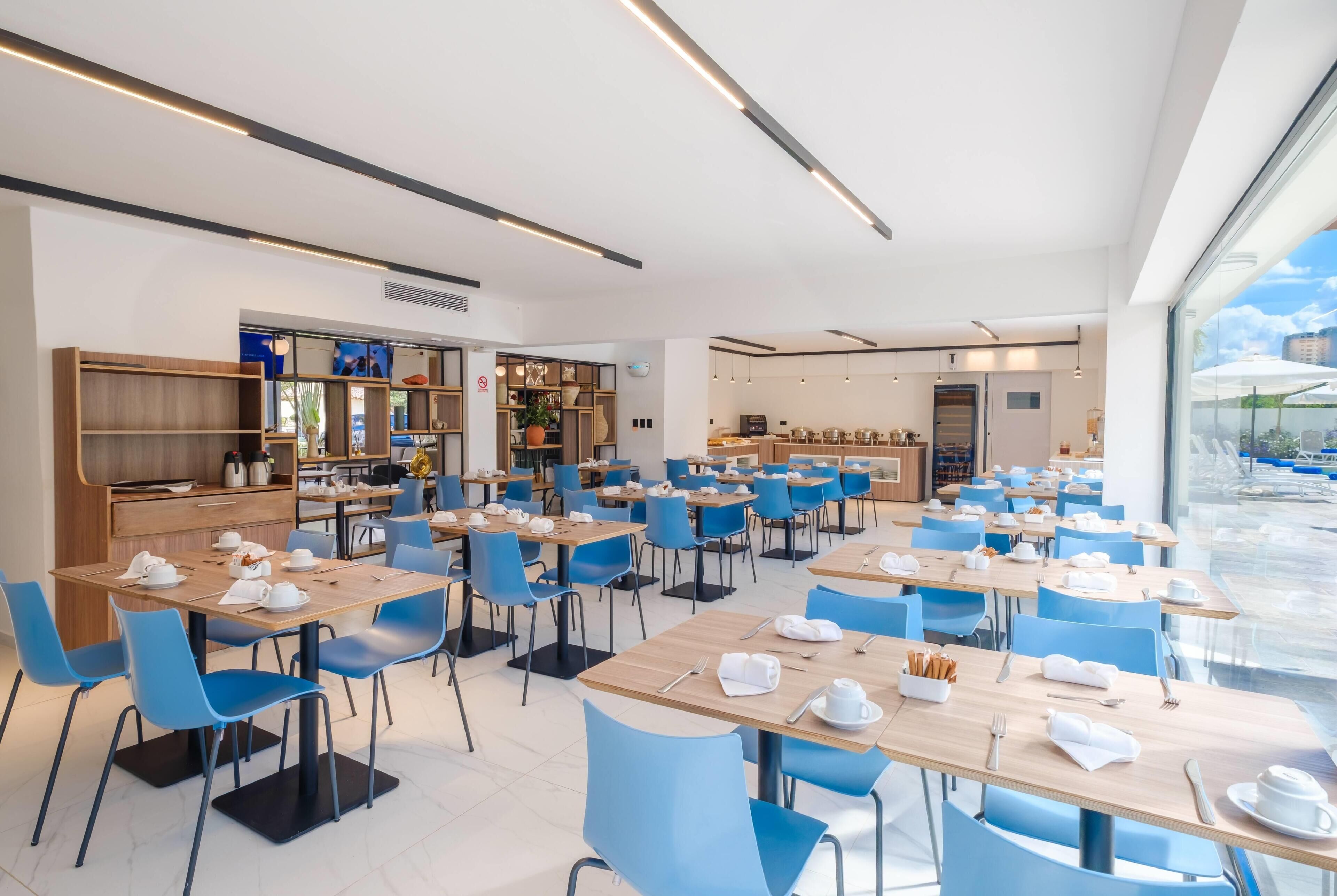 Uma área de refeições moderna, com mesas e cadeiras, uma cozinha ao fundo e vista através de grandes janelas.