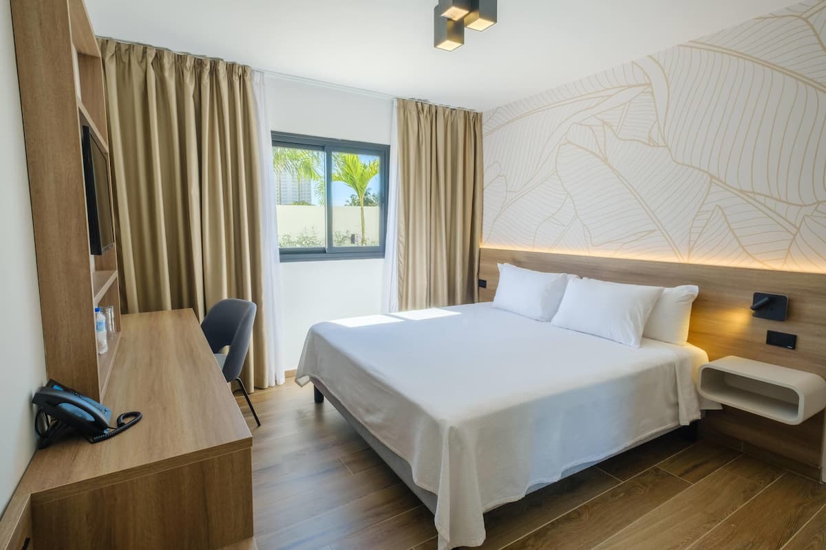 A hotel room with a bed, desk, chair, and a view of palm trees outside.
