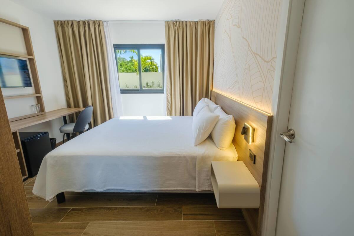 A hotel room with a bed, bedside table, desk, chair, and a window with curtains.