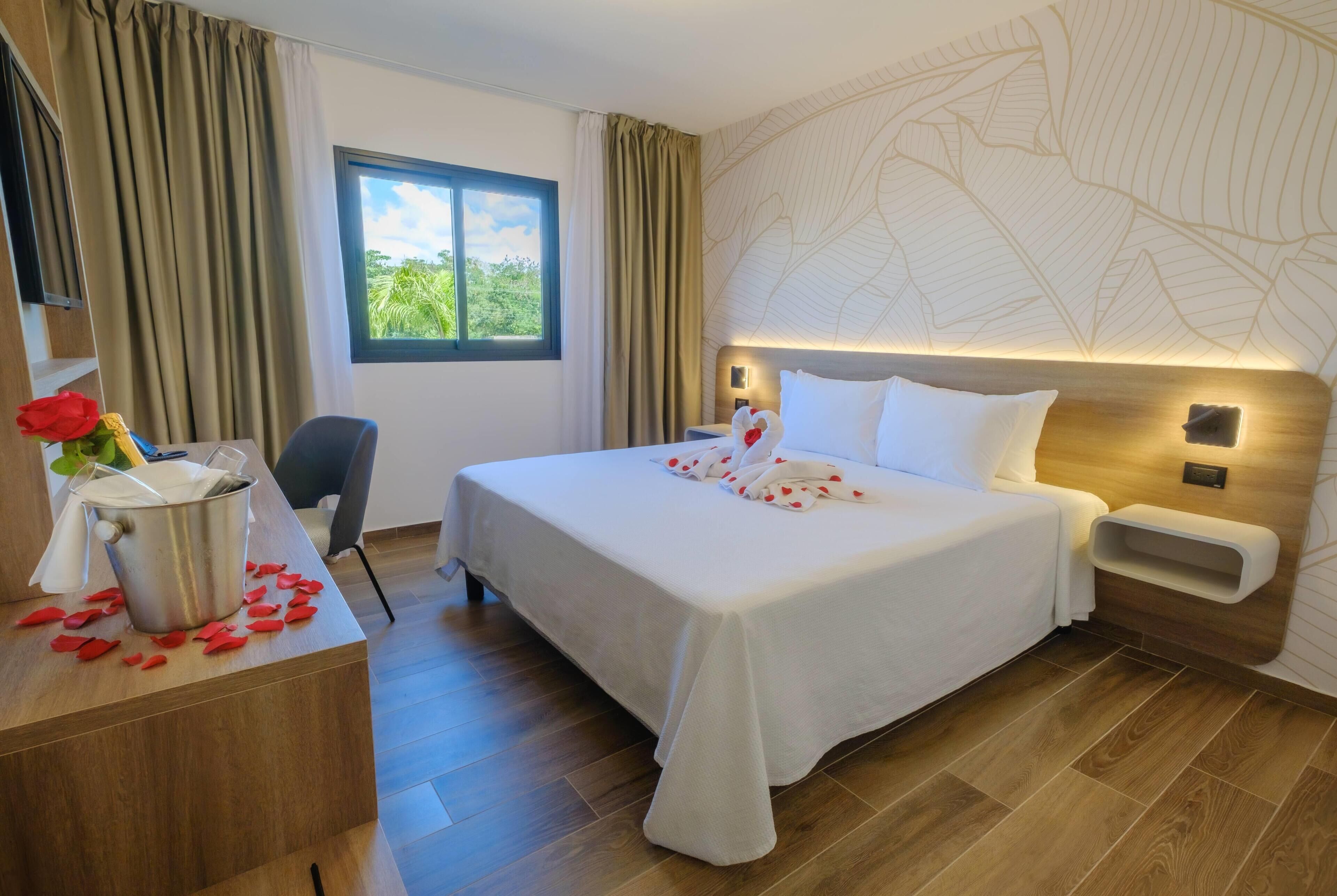 Une chambre d’hôtel avec un lit, un bureau sur lequel se trouve un seau rempli de pétales de rose, une chaise, une fenêtre avec des rideaux et un mur orné d’un motif de feuilles.