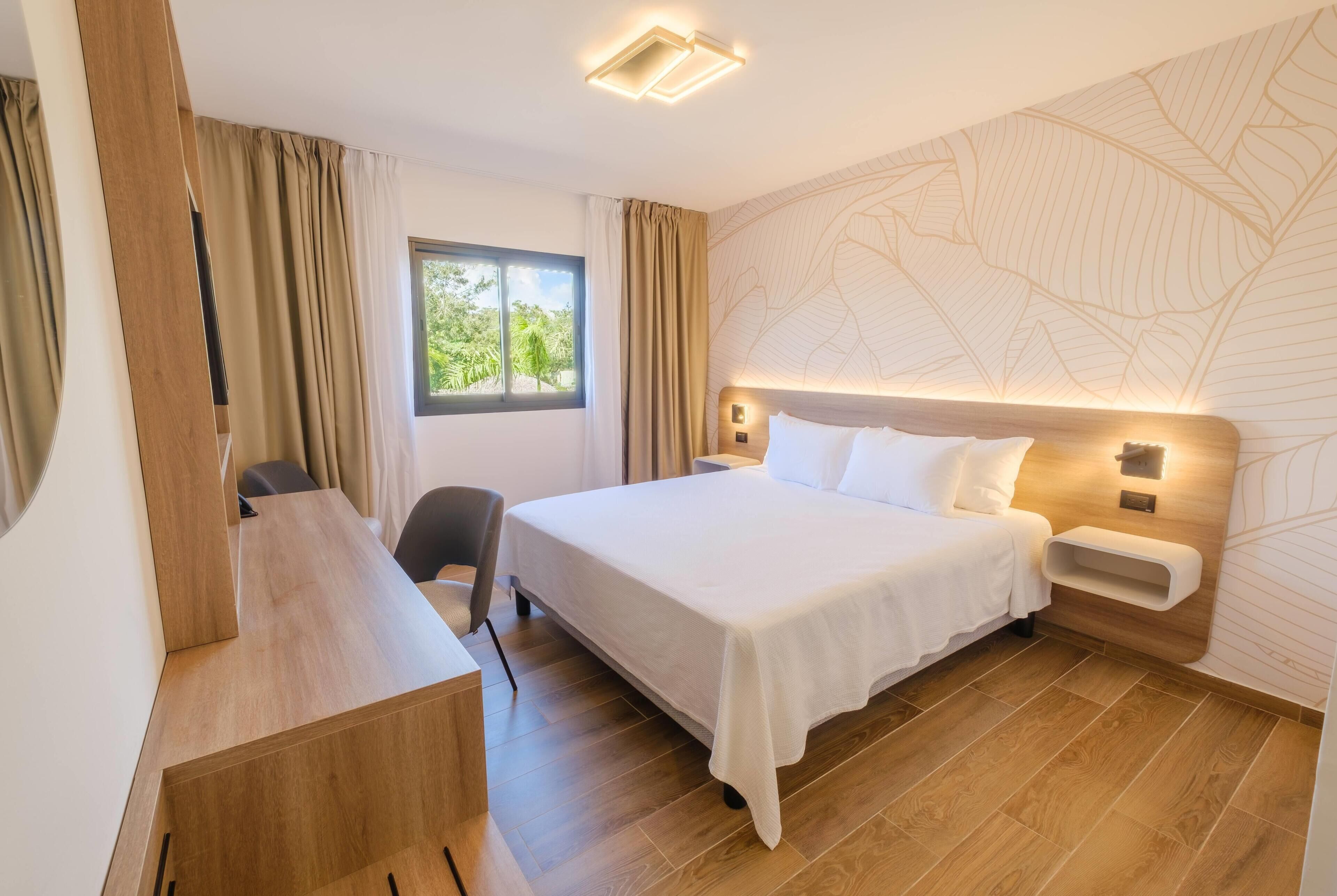 A hotel room with a bed, desk, chair, and a view of greenery through a window.
