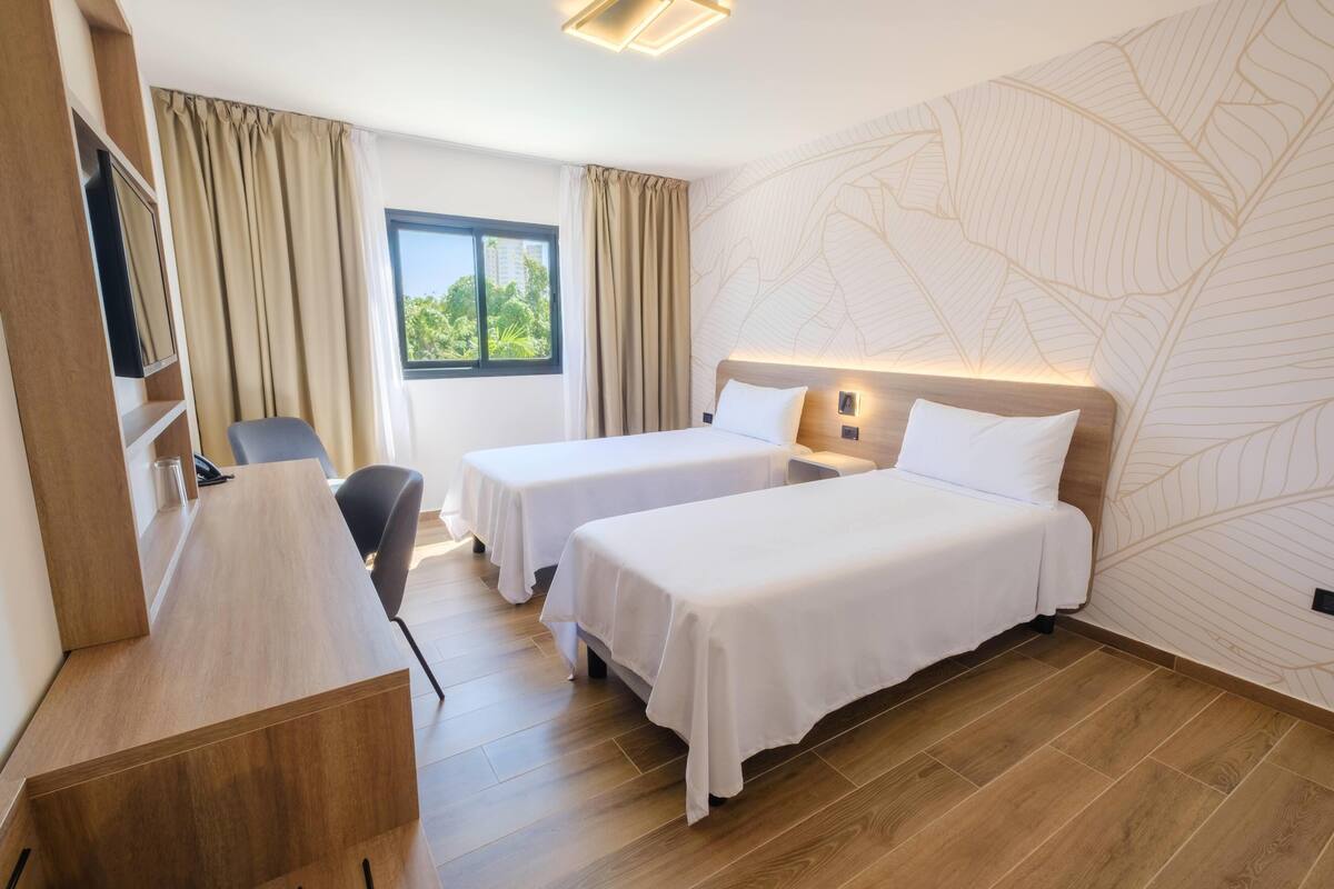 A hotel room with two beds, a desk, a chair, a TV, and a window with curtains.