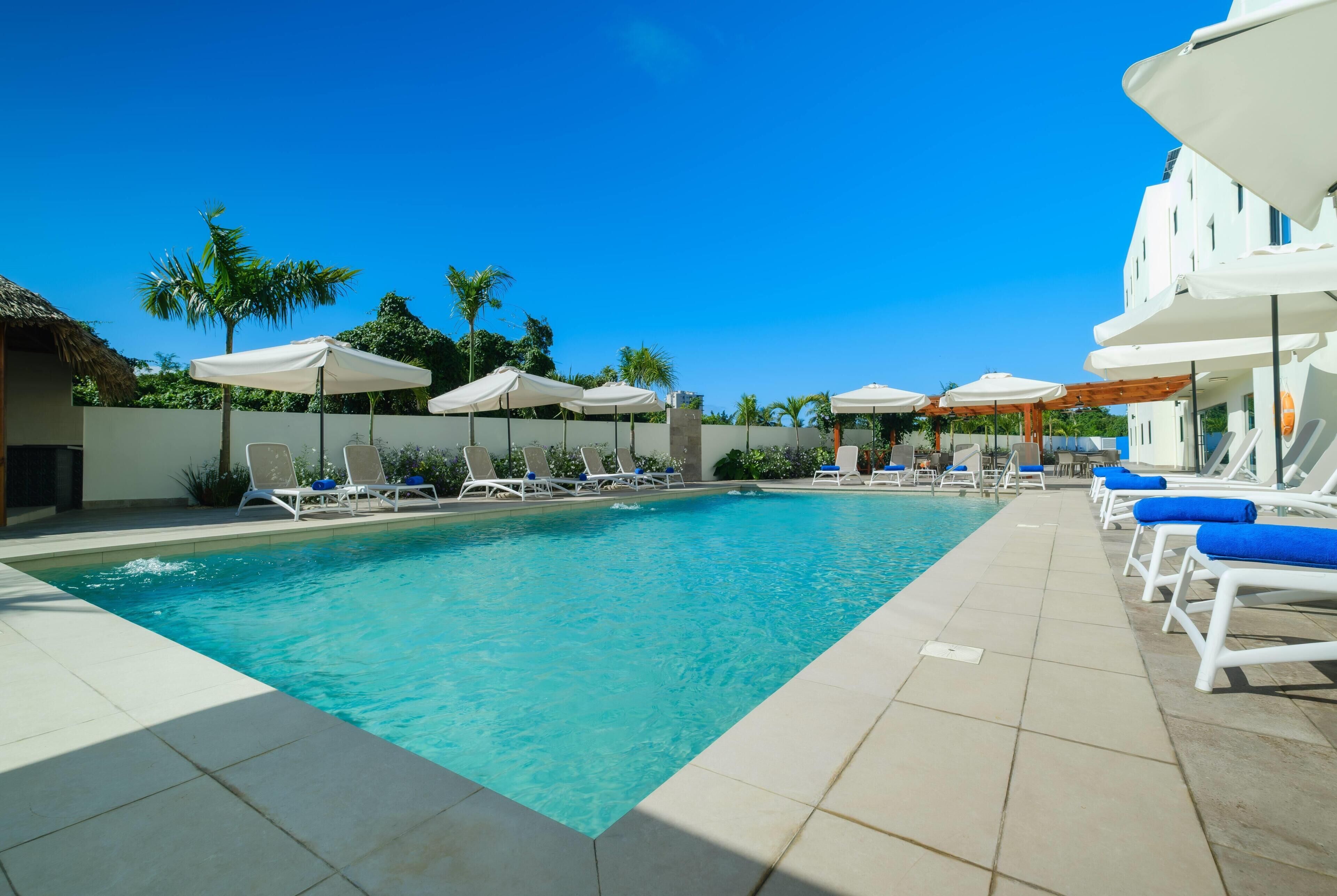 Ein Pool mit Liegestühlen und Sonnenschirmen, umgeben von Palmen und blauem Himmel.