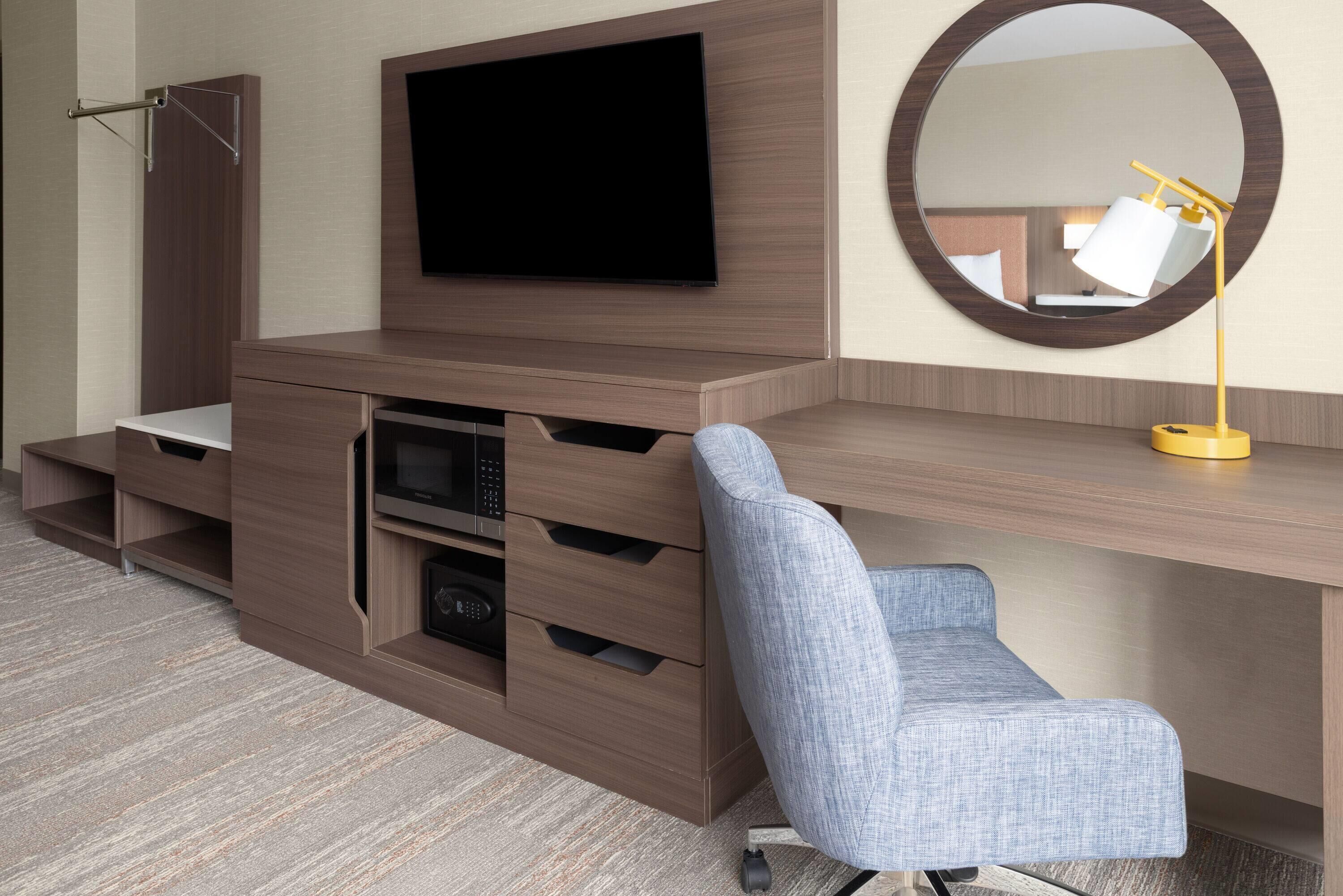 A modern room with a flat-screen TV mounted on a wooden cabinet, a round mirror on the wall, and a desk with a chair.