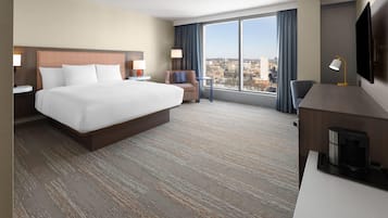 A hotel room with a large bed, a desk with a coffee maker, a TV, and a city view through the window.
