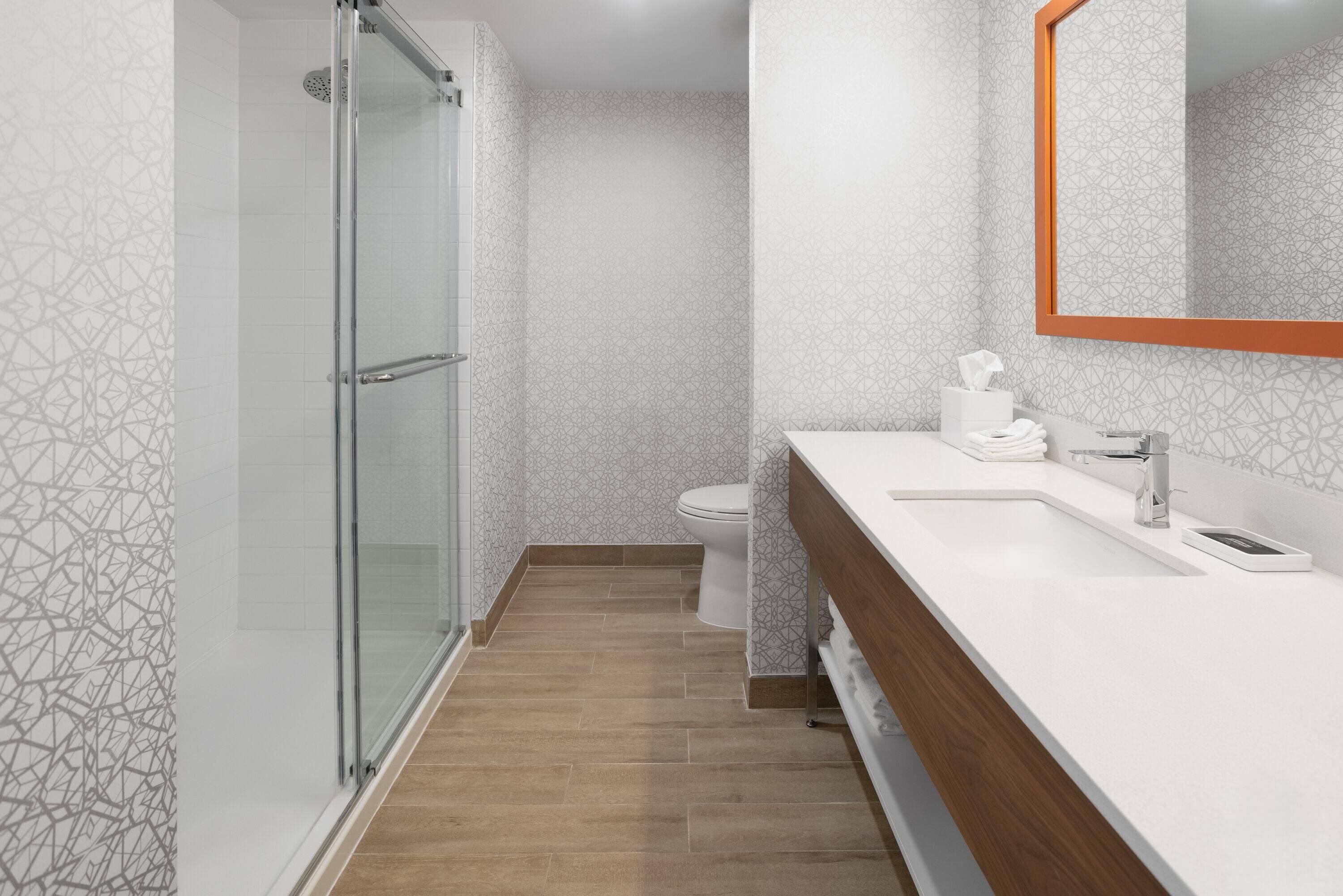 Un baño moderno con cabina de ducha de vidrio, un lavabo blanco con encimera de madera y un espejo grande con marco naranja.
