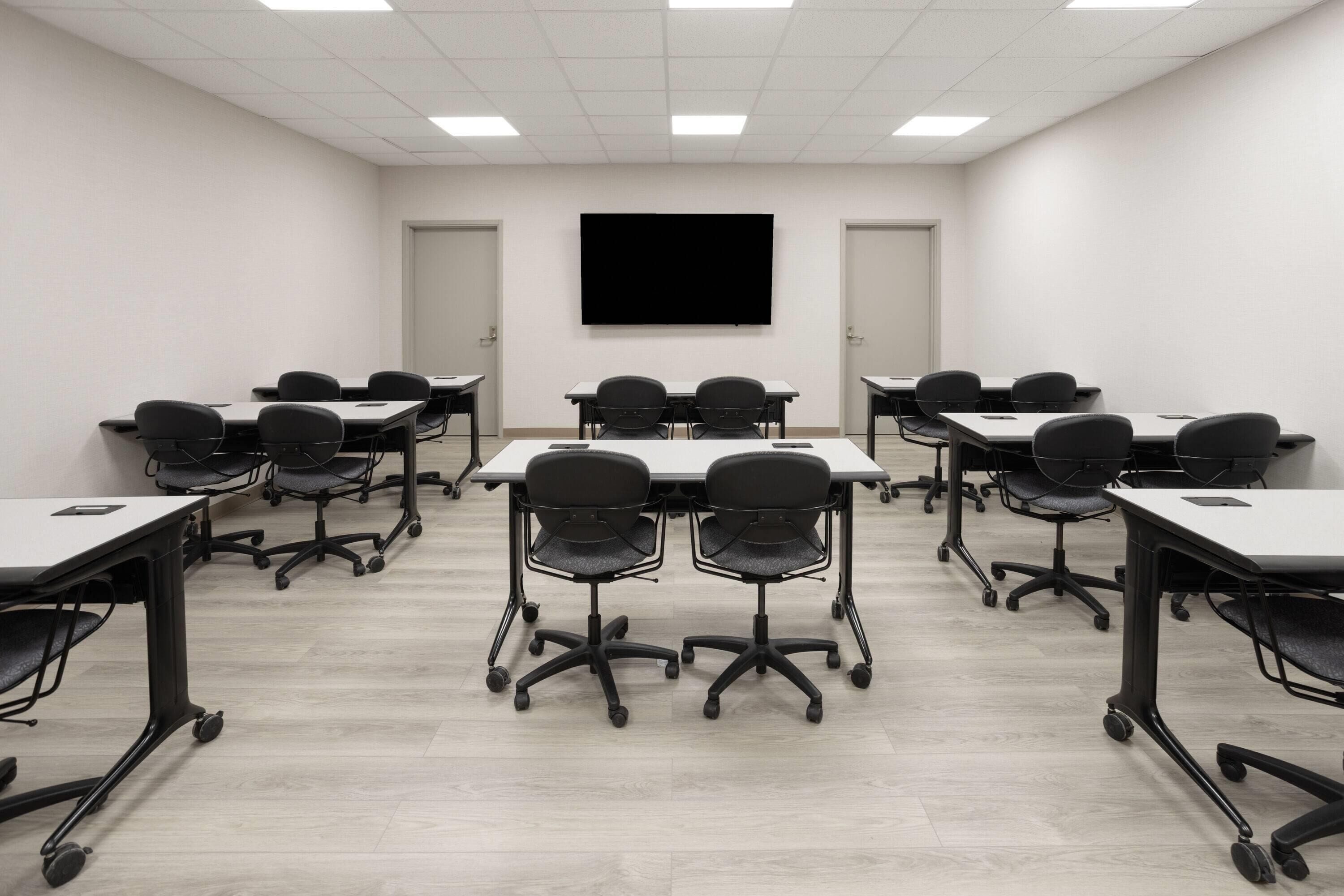 A conference room with a large flat-screen TV at the front, multiple tables with chairs, and a light-colored wooden floor.