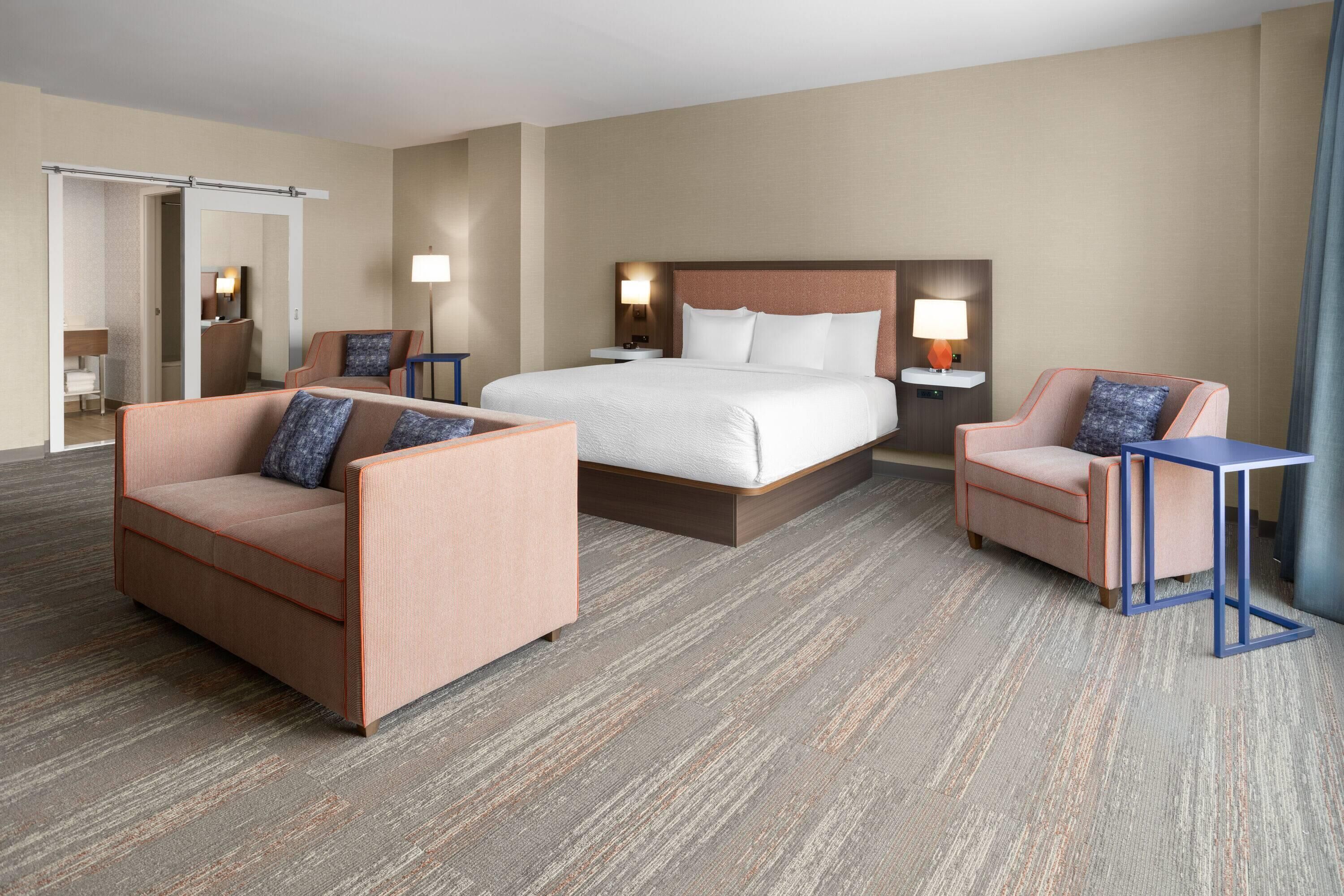 A hotel room with a large bed, two armchairs, a small table, and a mirror.