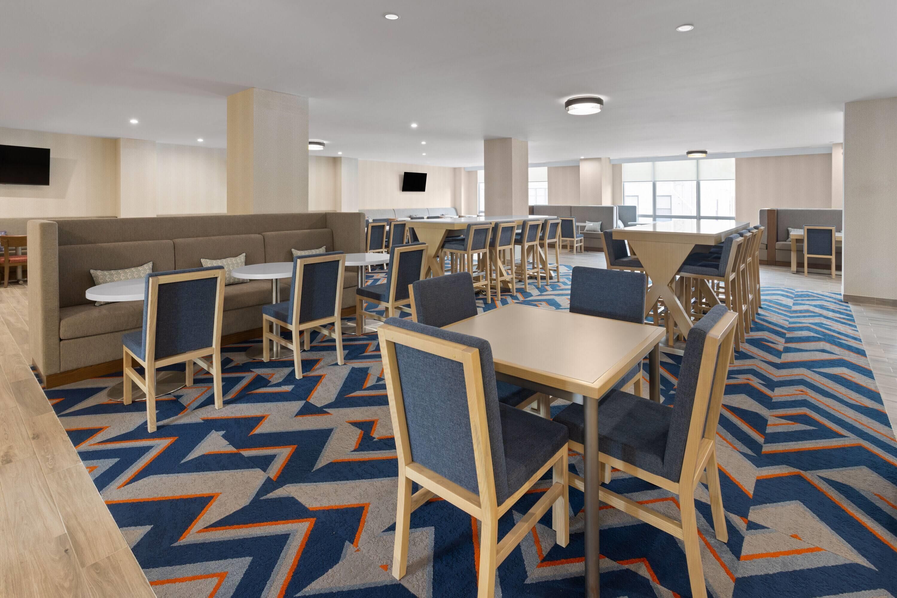 En modern hotellrestaurang med olika typer av sittplatser, inklusive bord och en soffa, ett mönstrat golv och en platt-TV som är monterad på väggen.