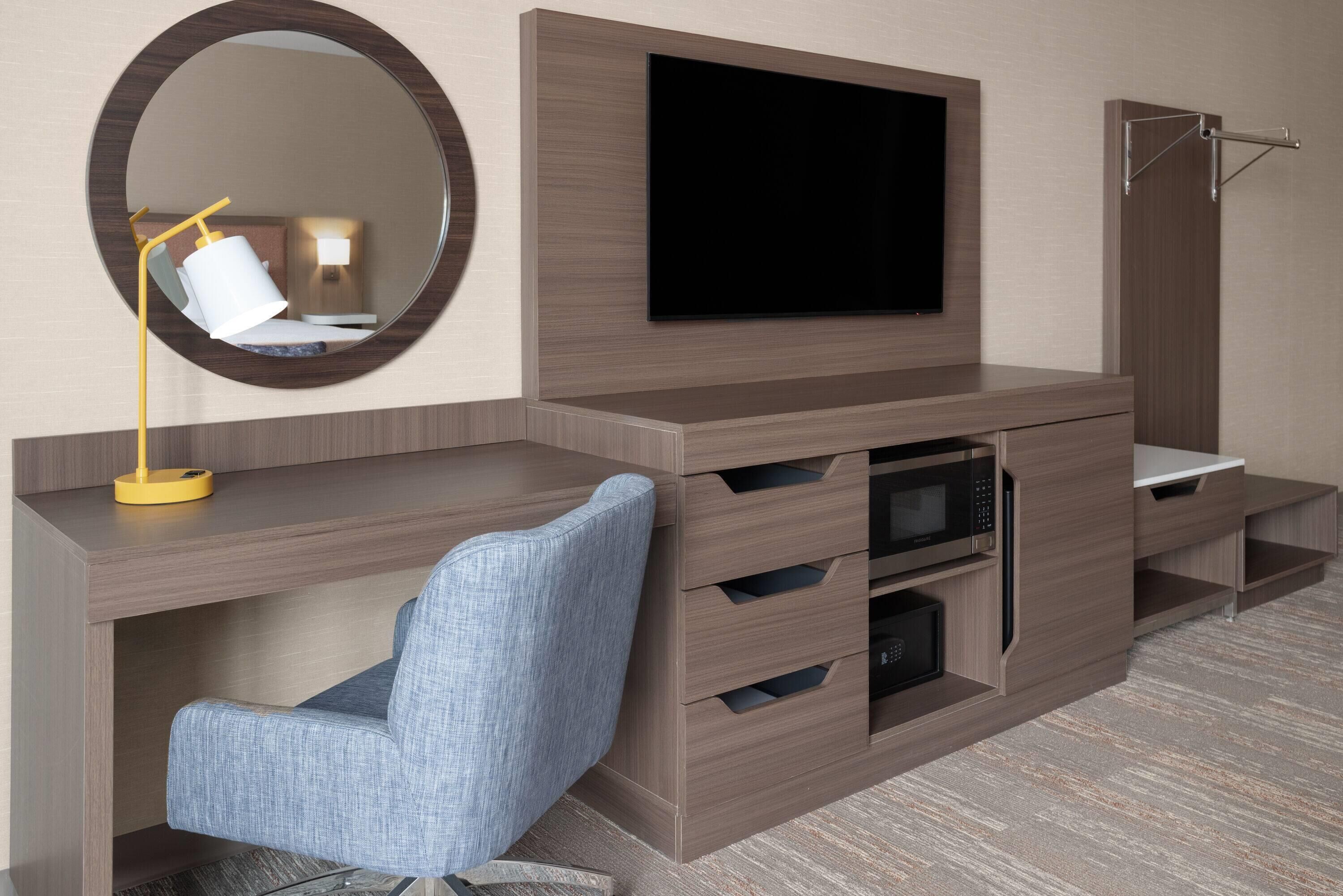 A modern hotel room with a flat-screen TV mounted on a wooden cabinet, a round mirror, a blue upholstered chair, and a built-in microwave.