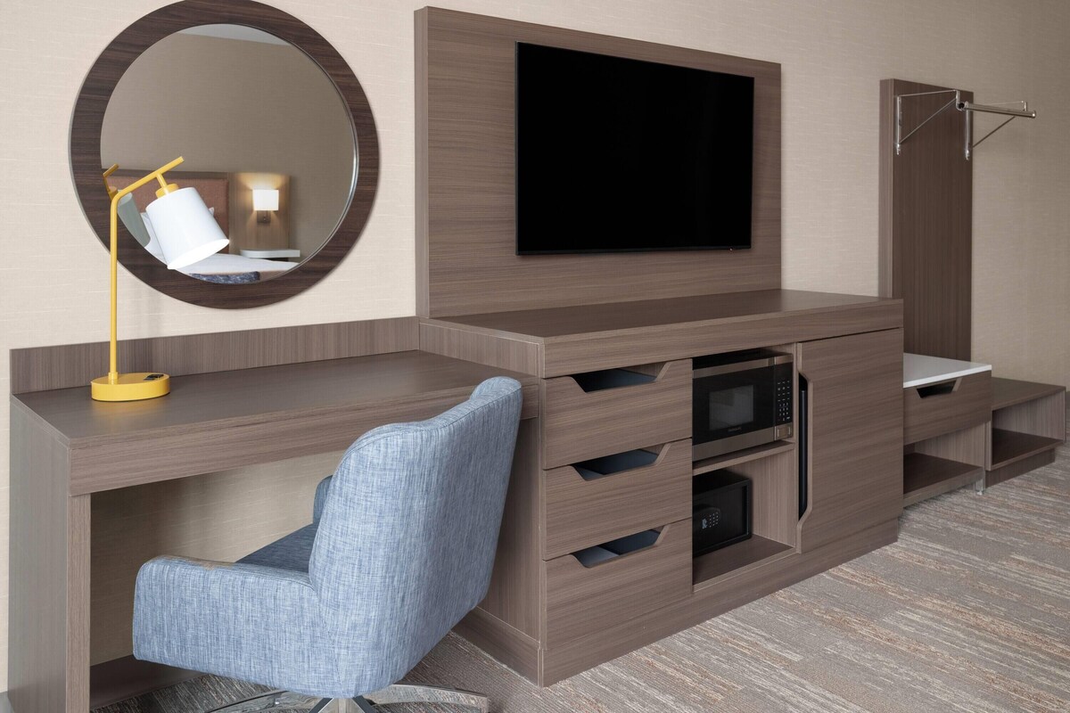A modern hotel room with a flat-screen TV mounted on a wooden cabinet, a round mirror, a blue upholstered chair, and a built-in microwave.