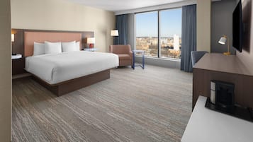 A hotel room with a large bed, a desk, a chair, a TV, and a view of the city.