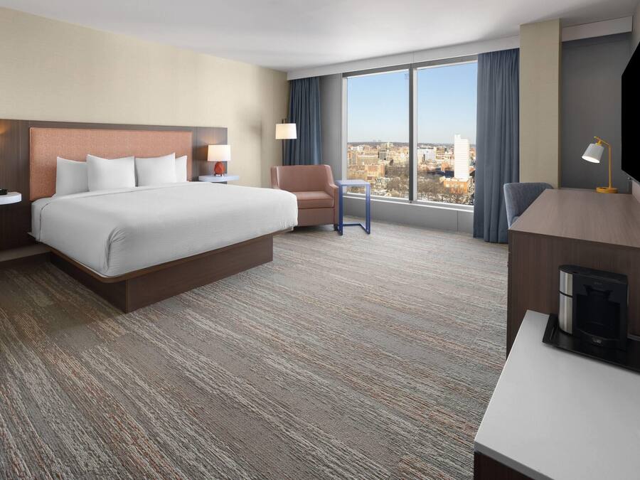 A hotel room with a large bed, a desk, a chair, a TV, and a view of the city.
