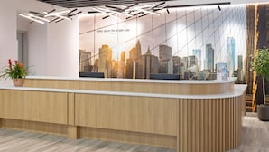 A reception desk with a cityscape mural, wooden paneling, and a potted plant. - La Quinta Inn & Suites By Wyndham Queens NYC/JFK AirTrain (Jamaica)
