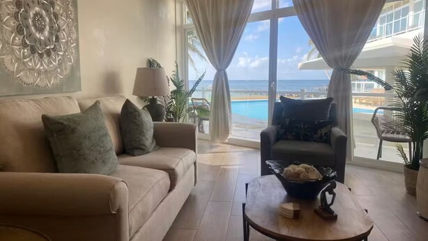 Ein Wohnzimmer mit einem beige farbenen Sofa, einem Couchtisch mit Dekorationselementen, Meerblick durch große Fenster und einer tropischen Pflanze.