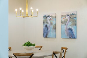 A dining area with a round table, chairs, a chandelier, and two wall-mounted paintings of a heron. - Styles Beach 1B - Beach View Condo w/ Pool (San Pedro)