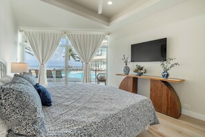 A bedroom with a large bed, a TV mounted on the wall, and a view of a pool and palm trees. - Styles Beach 1B - Beach View Condo w/ Pool (San Pedro)