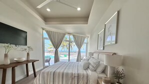 Un dormitorio con una cama grande, una mesita de noche de madera, un televisor montado en la pared y vista al mar a través de la ventana.