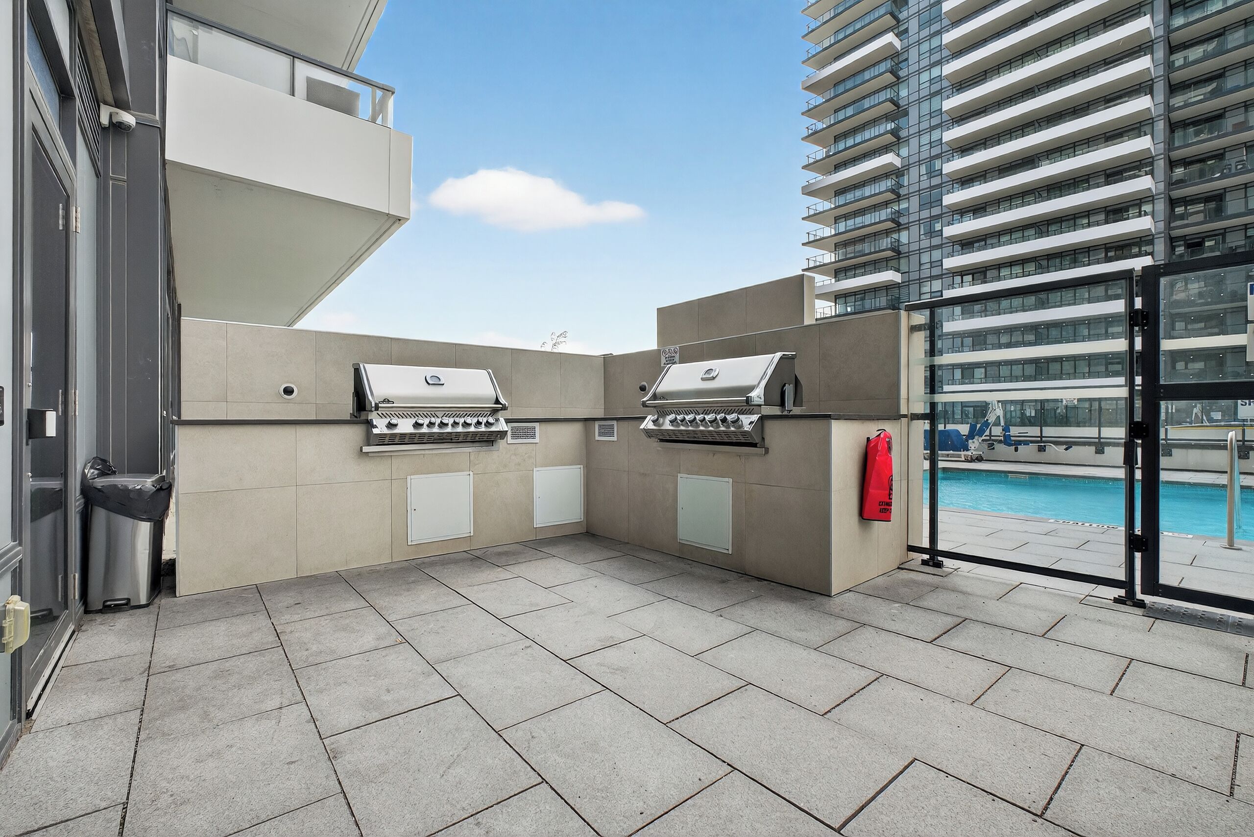 Un área de cocina al aire libre con dos parrillas, un contenedor de basura y vista a una piscina.