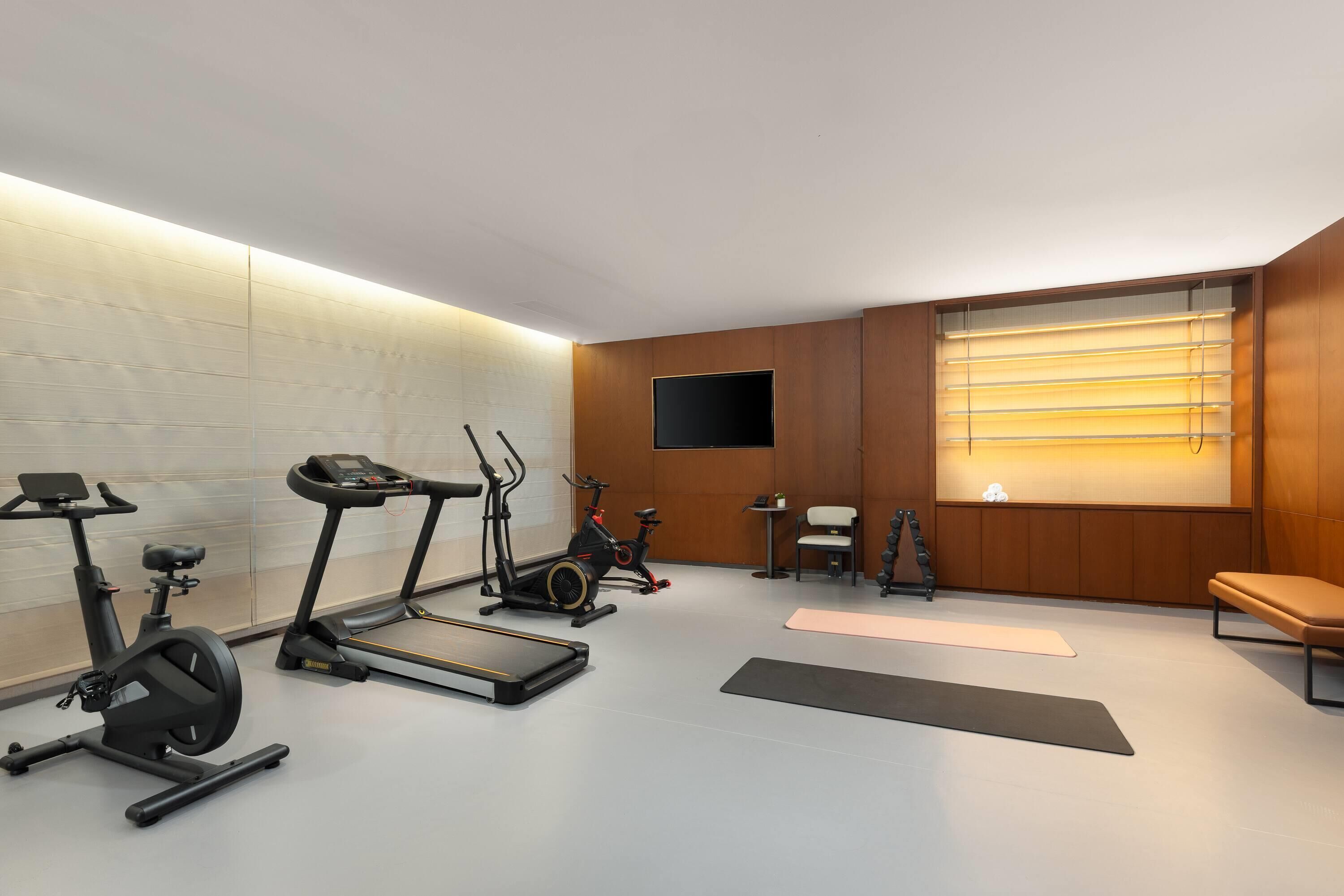 A modern gym with exercise equipment, a large window with blinds, and a bench.