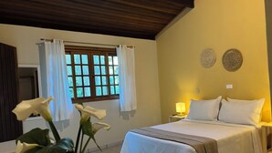 A bedroom with a bed, a nightstand, a lamp, a window with curtains, and a wall with two decorative plates. - Pousada Roca (Ibiúna)