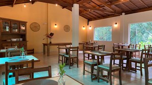A restaurant with wooden tables and chairs, a kitchen area, and large windows with a view of trees. - Pousada Roca (Ibiúna)
