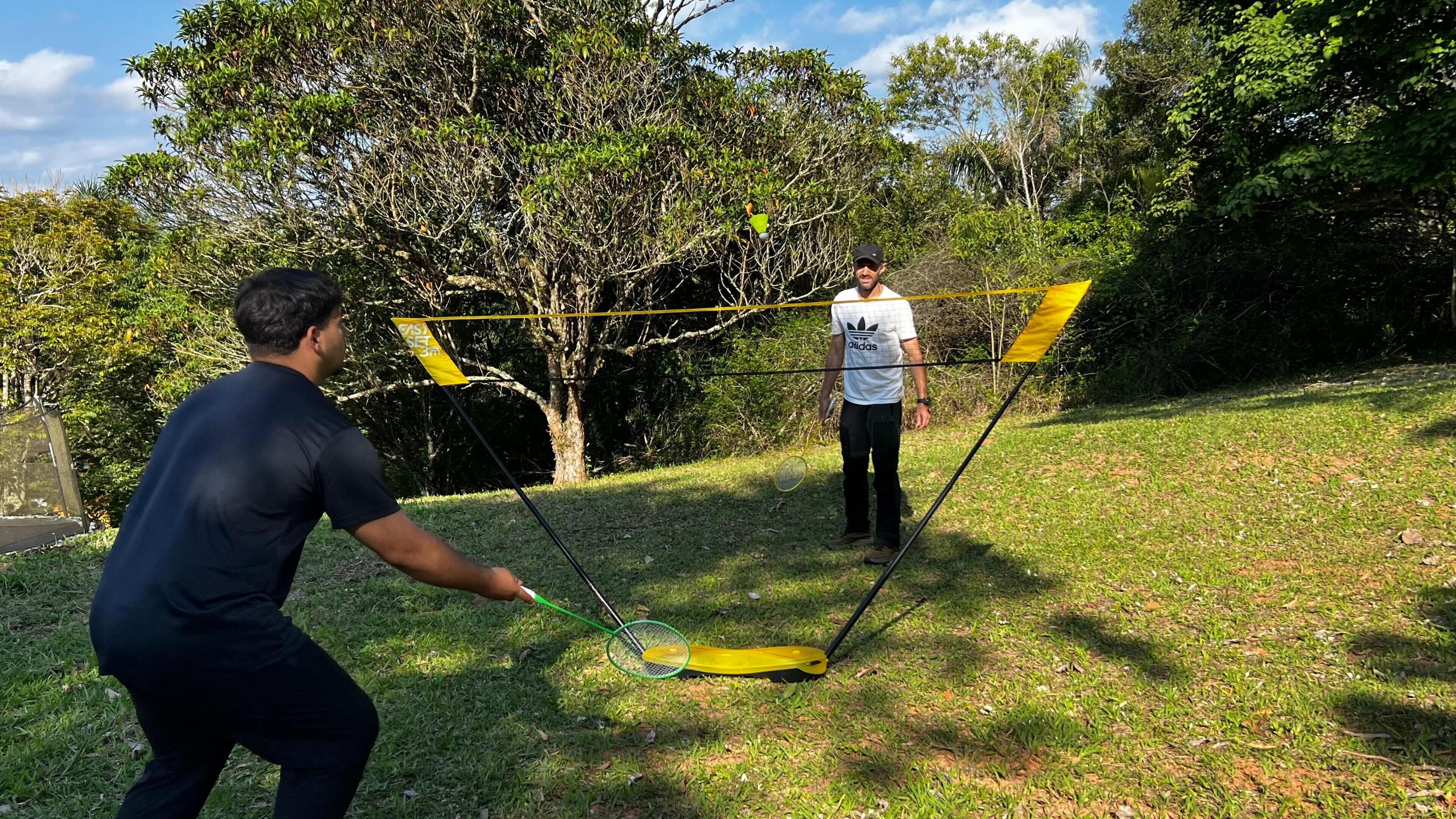 A person playing badminton on a grassy area with trees and a net in the background.