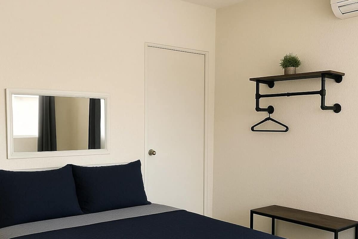 Un lit bien fait, recouvert d’une couvre-lit bleu foncé, une porte blanche, une petite table et une étagère murale agrémentée d’une plante et d’un porte-manteau.