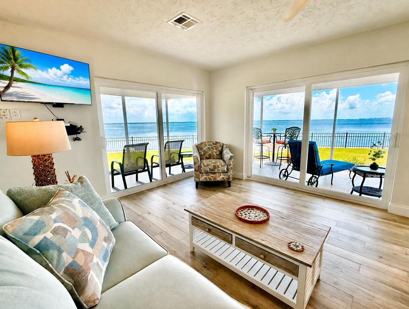 A living room with a sofa, armchair, and coffee table, large windows with a view of the beach, and a painting of a beach scene.