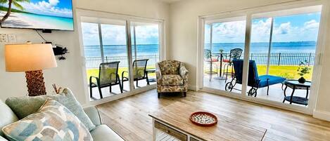A living room with a sofa, armchair, and coffee table, large windows with a view of the beach, and a painting of a beach scene.