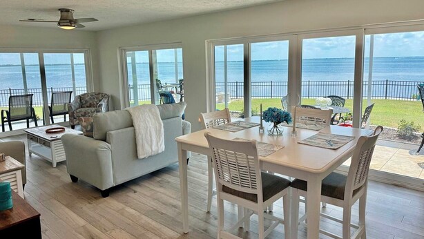 Een ruime woonkamer met een groot raam met uitzicht op zee, een plafondventilator en een eethoek met een tafel en stoelen.