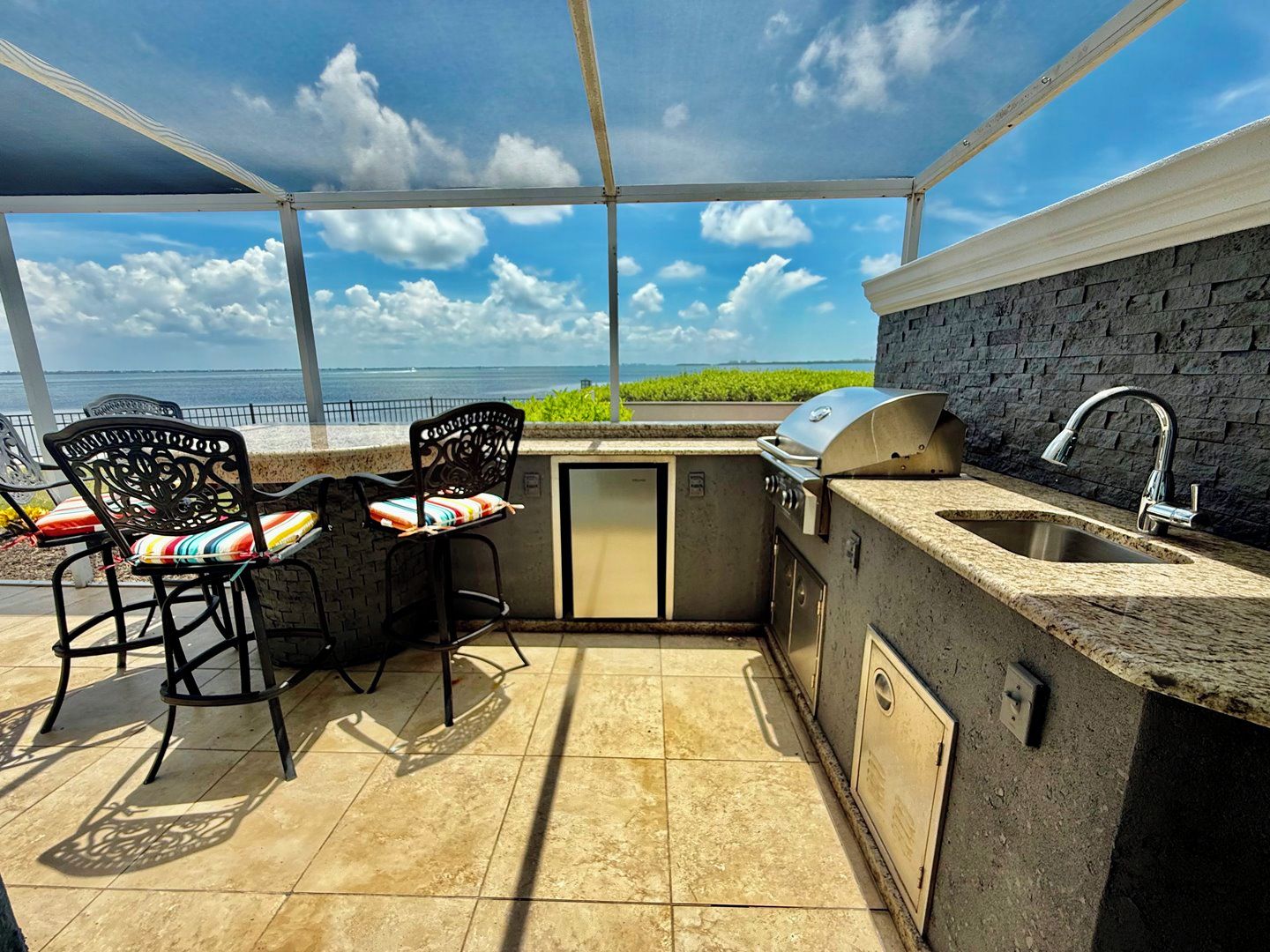 Une cuisine extérieure avec vue sur l’océan, un barbecue, un évier et deux chaises avec des coussins.
