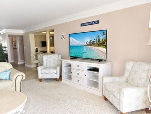 Un salon doté d’un téléviseur à écran plat, de deux fauteuils à motifs, d’un meuble de rangement blanc et d’un coin cuisine à l’arrière-plan.