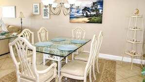 A dining area with a glass-top table, white upholstered chairs, a chandelier, and a painting of a beach scene. - Beachfront Vacation Rental With Full Gulf View (Longboat Key)