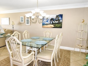 A dining area with a glass-top table, white upholstered chairs, a chandelier, and a painting of a beach scene. - Beachfront Vacation Rental With Full Gulf View (Longboat Key)