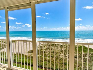 Une vue depuis un balcon donnant sur une plage, avec l’océan et une zone herbeuse.
