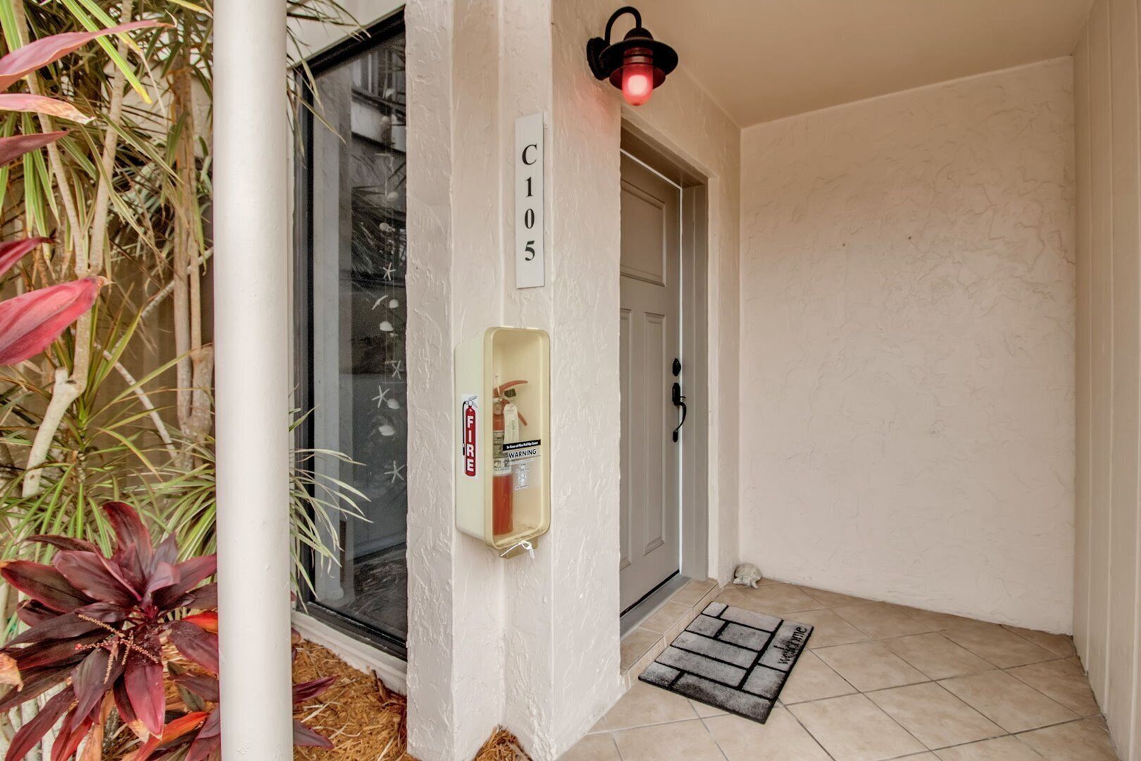 A clean, white-walled entrance with a fire extinguisher mounted on the wall, a door with a number sign, and a small mat at the entrance.