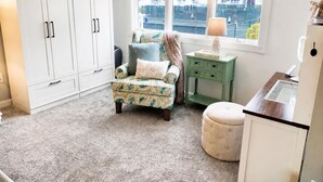 Quarto com poltrona estampada, uma mesinha com abajur, um armário branco e vista para edifícios pela janela.