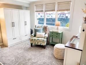 A room with a patterned armchair, a small table with a lamp, a white cabinet, and a view of buildings through the window. - Gorgeous 3-bedroom beachfront unit at Club Longboat! (Longboat Key)