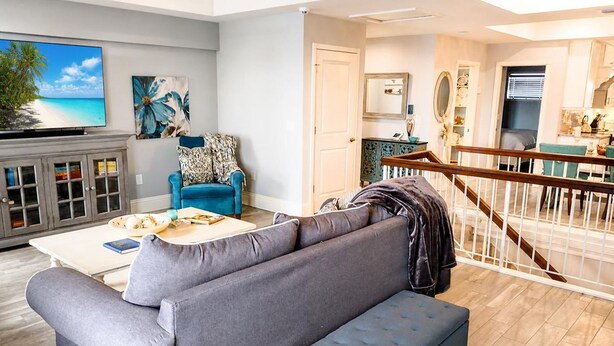 Sala de estar com sofá cinza, poltrona azul e uma mesa de centro. Escada leva a outro cômodo.