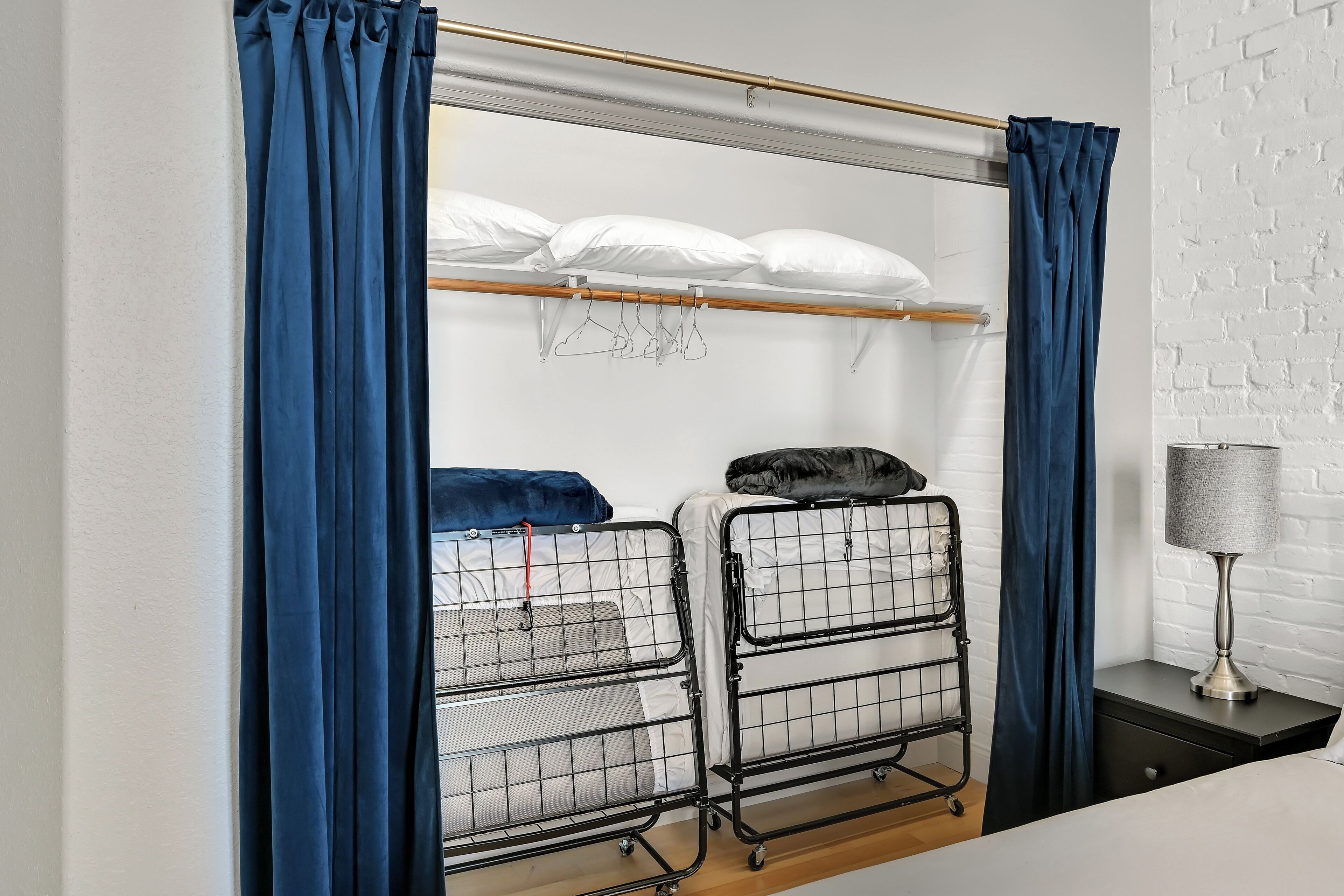 A bunk bed with a metal frame, white bedding, and a black pillow. There is a wooden shelf above the bed with white pillows. A lamp is on a bedside table next to the bed.