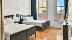 A room with two beds, a white brick wall, and large windows with curtains.