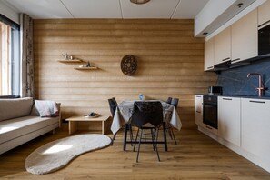 A modern kitchen with wooden cabinetry, a dining area with a table and chairs, and a sofa with a view of the outdoors. - Le Chalet Lingonberry D7 by Aavalevi (Levi)