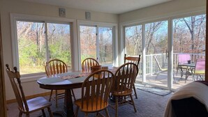 Un espace repas avec une table ronde et des chaises en bois, de grandes fenêtres donnant sur la verdure et un coin salon extérieur abrité.