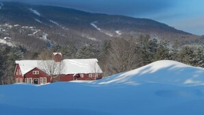 Ein rotes Bauernhaus mit schneebedecktem Dach, umgeben von verschneiter Landschaft und fernen Bergen.