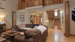 Ein geräumiger Wohnbereich mit braunem Sofa, einer Holztreppe und einer hohen Decke mit sichtbaren Holzbalken.